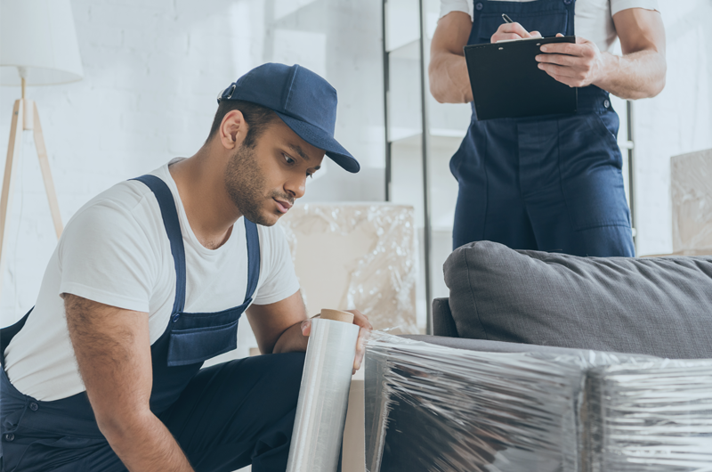 Professional mover in blue overalls wrapping a sofa to move.